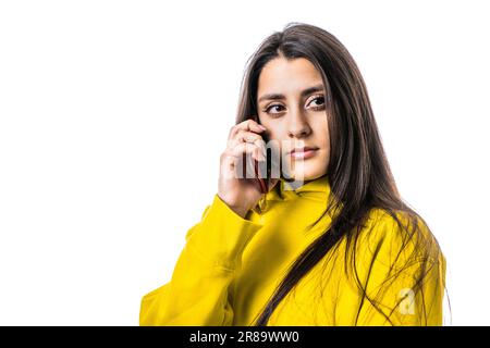 Ritratto in studio di bella ragazza adolescente caucasica millenaria posa in felpa gialla con cappuccio, chattando a smartphone con sfondo bianco luminoso. S Foto Stock