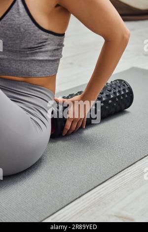 Vista ritagliata della giovane donna sfocata in abbigliamento sportivo che tocca il moderno massaggiatore a rulli e che si siede sul tappetino fitness a casa, senso di tranquillità e promuovere Foto Stock