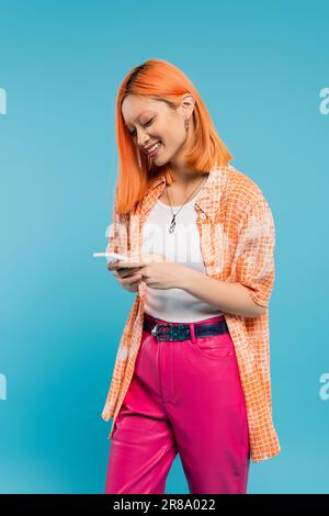social networking, allegra donna asiatica con messaggi colorati sui capelli, utilizzando lo smartphone, in piedi su sfondo blu, sorridente, camicia arancione, abbigliamento casual, Foto Stock