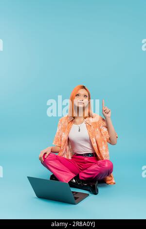 pensiero creativo, idea gesto, soluzione, rosso donna asiatica in camicia arancione seduta con gambe incrociate vicino al portatile, guardando verso l'alto e puntando con pinna Foto Stock