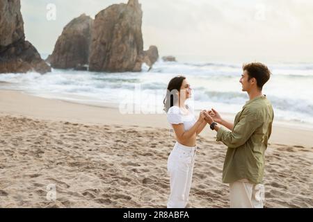 Coppia amorevole tenendo le mani e guardando l'un l'altro, avendo data romantica sulla spiaggia vicino alla riva dell'oceano, spazio libero. Foto Stock