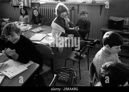 Scuola media, scolari e scolaresche in classe. Studentessa disabile che indossa calibri o ha chiamato rinforzi per le gambe 1970S. Non riesce a girare facilmente sulla sua sedia per vedere l'insegnante che sta parlando con la classe, quindi le è permesso di sedersi sul tavolo. Le bretelle sono semplicemente visibili sedute sulla scrivania condivisa. Una borsa per la Scozia per la Coppa del mondo di Monaco 1974 è sul pavimento. Breasclete, vicino Carloway Isola di Lewis e Harris, Ebridi esterne, Scozia circa giugno 1974. HOMER SYKES Foto Stock