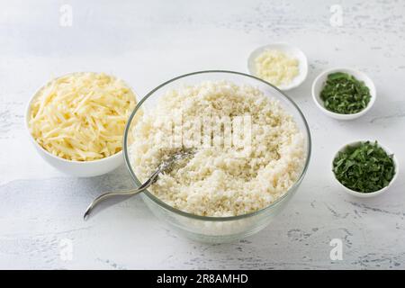 Cavolfiore grattugiato, riso cavolfiore o couscous cavolfiore circondato da erbe tritate, aglio e formaggio grattugiato su un tavolo azzurro. Cucina Foto Stock