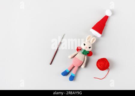 Coniglio giocattolo lavorato a maglia in un cappello rosso e sciarpa con palla di filo e gancio a maglia su sfondo grigio. Articoli e accessori per bambini. Omaggio per le vacanze invernali Foto Stock
