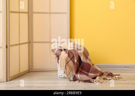 Cestino con nuova coperta morbida e schermo pieghevole vicino alla parete gialla in camera Foto Stock