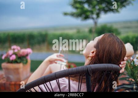 Attraente giovane donna è seduta sulla sedia di vimini nel cortile della casa. Una ragazza affascinante sogna, sorride e beve vino o cocktail in terrazza. P Foto Stock