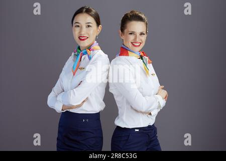Ritratto di felici e moderni assistenti di volo femminili in gonna blu, camicia bianca e sciarpa isolata su sfondo grigio. Foto Stock