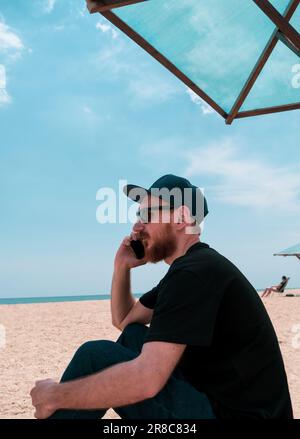 Uomo millenario beared in t-shirt nera snapback sul mare blu paesaggio con smartphone in mano parlando. Giovane adulto che chiama amici sul cellulare, Foto Stock