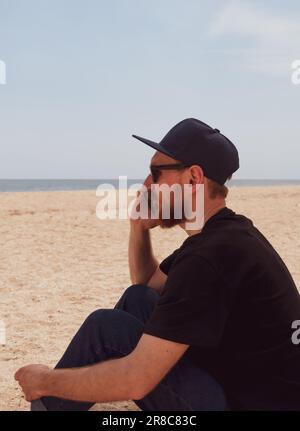 Uomo millenario beared in t-shirt nera snapback sul mare blu paesaggio con smartphone in mano parlando. Giovane adulto che chiama amici sul cellulare, Foto Stock