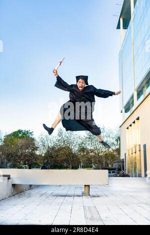 Felice uomo laureato in un abito nero saltando mentre tiene il suo diploma Foto Stock