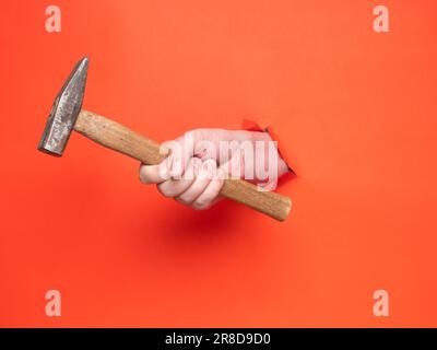 La mano di un uomo tiene un martello attraverso carta arancione strappata. Concetto, passare attraverso carta arancione strappata. Foto Stock