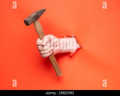 La mano di un uomo tiene un martello attraverso carta arancione strappata. Concetto, passare attraverso carta arancione strappata. Foto Stock