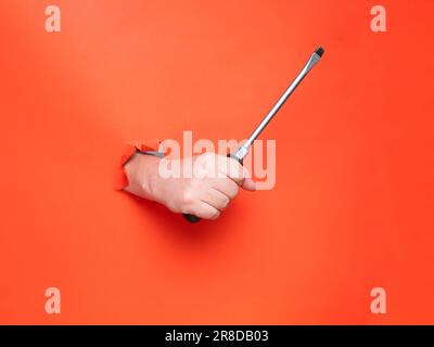 Tenere a mano un cacciavite grande attraverso carta arancione strappata. Niente faccia, concetto. Foto Stock