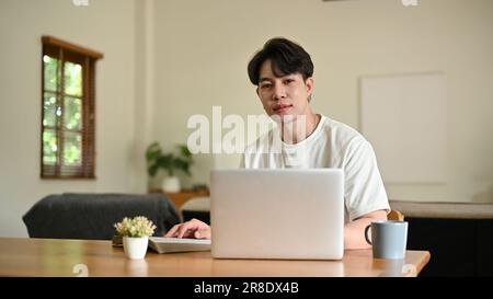 Un bel giovane asiatico in abiti casual siede a un tavolo con il suo laptop nel suo salotto minimalista. Foto Stock