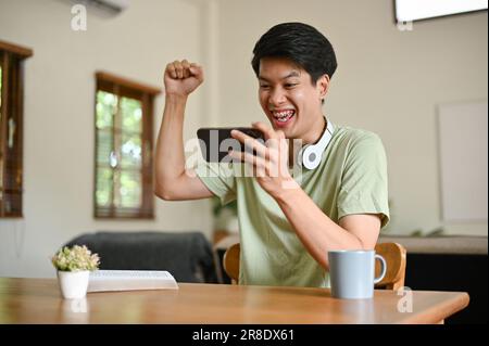 Un giovane asiatico eccitato e gioito in abiti comodi solleva la mano, festeggiando una partita sportiva o giocando un gioco per cellulare sul suo telefono Foto Stock