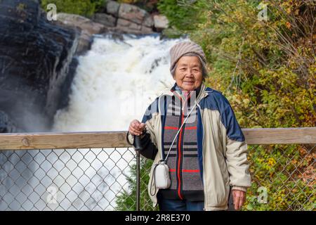 Turista cinese anziano in posa per le foto di fronte alle cascate al Canyon Sainte-Anne. Quebec in autunno. Canada. Foto Stock