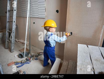 Tutta la lunghezza del lavoratore di costruzione del bambino che dipinge la parete con la spazzola nell'appartamento in fase di ristrutturazione. Bambino in casco di sicurezza e tute da lavoro giocando a casa, in piedi in stanza con gli attrezzi e la scala. Foto Stock