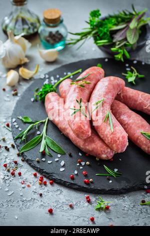 Salsicce crude su ardesia, con erbe e spezie, preparate per grigliate e barbecue Foto Stock