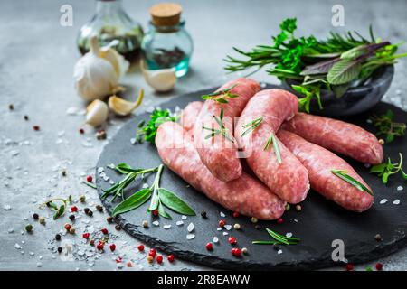 Salsicce crude su ardesia, con erbe e spezie, preparate per grigliate e barbecue Foto Stock