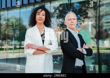 Ritratto di persone di affari a braccia incrociate che guardano la fotocamera con espressione di successo. Coppia di colleghi orgogliosi e multirazziali che si trovano in ufficio. Due partner esecutivi o colleghi sul posto di lavoro. Foto di alta qualità Foto Stock