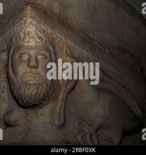 Testa umana accanto a un basilisk con denti nudi. Scolpito in marmo su capitale romanica nel chiostro aggiunto alla fine del 1200s alla chiesa abbaziale di San Michele, o église abbatiale St-Michel, a Saint-Génis-des Fontaines, Pyrénées-Orientales, Occitanie, Francia. Foto Stock