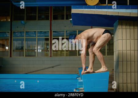 Atleta anziano in piedi sul blocco di partenza che si prepara a saltare in piscina Foto Stock