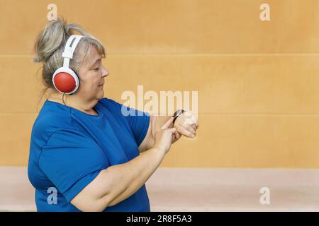 Donna più anziana con sovrappeso, abbigliamento sportivo e cuffie consulta il suo smartwatch mentre si allena nel parco. Foto Stock