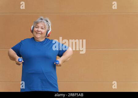 Donna obesa bianca più anziana che si esercita con manubri e cuffie ascoltando musica per la perdita di peso. Isolato su sfondo arancione Foto Stock