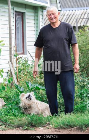 Rilassamento uomo anziano rimanere al sole al bordo del suo giardino con il suo bel cane bianco outbred godendo l'estate come pomeriggio di agosto. Foto Stock