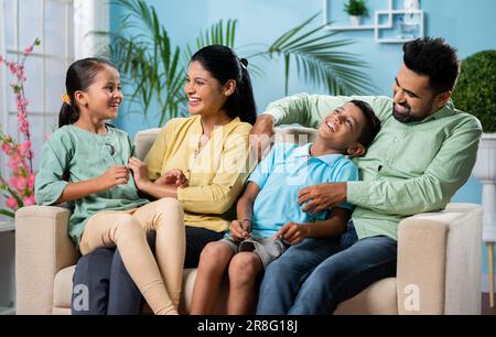 Concentratevi sulla madre, la felice coppia indiana con i bambini che giocano coccolandosi o facendo il solletico mentre si siedono seduti sul divano a casa - concetto di legame familiare Foto Stock
