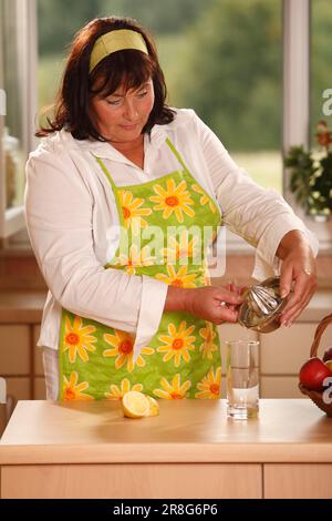 La donna versa il succo di limone in un bicchiere d'acqua, spremiagrumi, fascia per capelli, spremitura, spremitura fresca, versamenti, versamenti, versamenti Foto Stock