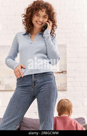 giovane donna allegra che parla su smartphone in piedi vicino al bambino, equilibrio tra lavoro e vita, genitorialità moderna, lavoro remoto, carriera e famil Foto Stock