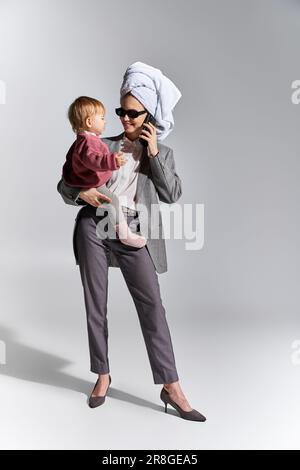 multitasking, donna in occhiali da sole che tiene in braccio figlia e in piedi con asciugamano sulla testa, stile di vita di equilibrio, donna d'affari in abbigliamento formale parlando Foto Stock