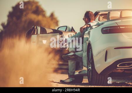 Uomo di mezza età di successo e ricco seduto nel suo moderno bianco convertibile godendo colorato tramonto. Vita felice. Foto Stock