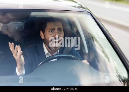 Il driver maschio arrabbiato dell'automobile grida ad altri driver e pedoni che ostruiscono il traffico, uomo d'affari adulto maturo in un vestito di affari è in ritardo per una riunione di affari in un automobile. Foto Stock