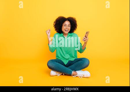 Foto a tutta lunghezza di entusiasta ragazza riccia afro-americana o brasiliana, siede su sfondo giallo isolato, utilizza smartphone, navigare in Internet, si rallegra delle notizie, guarda la fotocamera in stupore Foto Stock