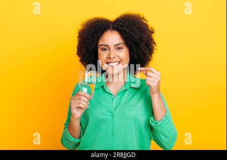 Assunzione giornaliera di vitamine e integratore. Felice bella donna brasiliana o afro-americana in camicia casual, tenendo la pillola e il bicchiere d'acqua, guarda la macchina fotografica, sorriso, stand su sfondo giallo isolato Foto Stock