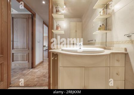 Piccolo bagno con lavabo in porcellana bianca incorporato in un piano di marmo, un piccolo armadio in legno bianco con uno specchio integrato e una porta in quercia e. Foto Stock