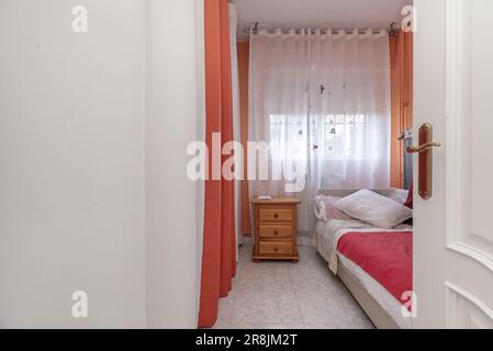 Ingresso ad una camera da letto con una porta in legno laccato bianco, un armadio con tende bianche e rosse e un letto singolo con un tavolino in legno di pino Foto Stock