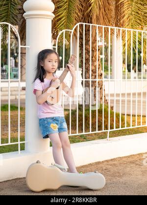 Bambina asiatica giocare l'ukulele, nel giardino, appoggiandosi contro il recinto, pratica per giocare Foto Stock