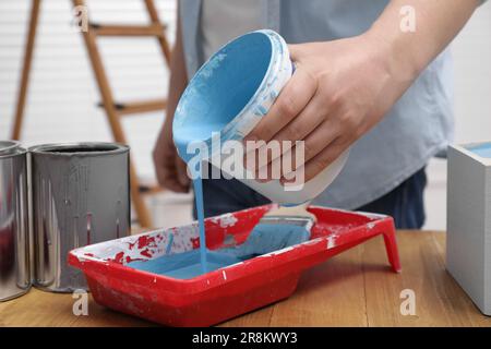 Uomo che versa la vernice blu chiaro dal secchio nel vassoio al tavolo di legno all'interno, primo piano Foto Stock