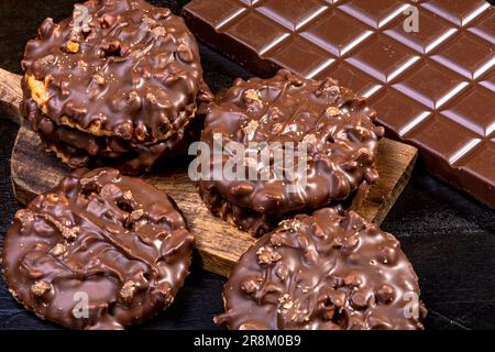 Biscotti ricoperti di cioccolato e nocciola su un vassoio di legno, accanto ad una tavoletta di cioccolato al latte, isolati su un fondo scuro dipinto a mano dalla fotogra Foto Stock