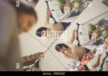 Vista da un angolo basso della donna con carrello della spesa che puzza di frutta e si riflette nello specchio del supermercato Foto Stock