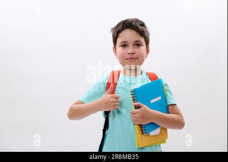 Bambino della scuola intelligente, bel ragazzo della preda sorride guardando la macchina fotografica, sborsando, mostrando il cartello di approvazione su sfondo bianco Foto Stock