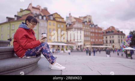 Signora anziana siede sui gradini bevendo caffè e utilizzando uno smartphone nel centro storico di una vecchia città europea. Piazza del Palazzo, Città Vecchia di Varsavia Foto Stock