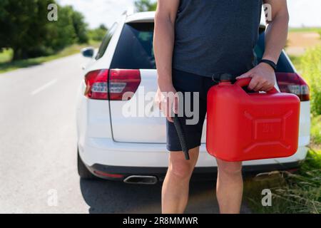 Un uomo tiene in mano una lattina rossa accanto a un'auto bianca. Fermarsi per fare rifornimento, problemi sulla strada con il rifornimento Foto Stock