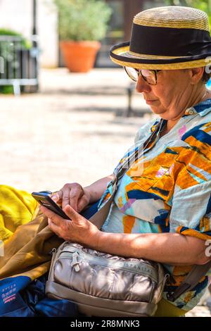 Donna anziana matura in cappello di paglia sul centro città panca giardino toccando messaggio sul telefono cellulare - Tours, Indre-et-Loire (37), Francia. Foto Stock