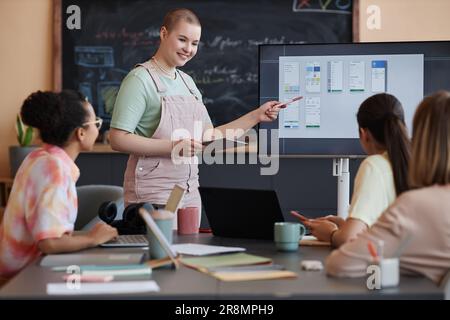 Ritratto di donna sorridente calva che indica lo schermo del computer con un progetto di design, mentre si presenta a tutta la squadra femminile in ufficio Foto Stock