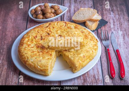 Tipica frittata spagnola di patate preparata sul momento su un tavolo di legno con posate, pane e olive Foto Stock