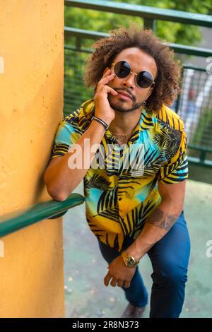 Passato di un uomo attraente con capelli afro in una camicia estiva di palma su uno sfondo giallo e una scala Foto Stock